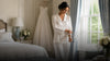 Bride wearing elegant button-up pajamas in a bright bridal suite with her wedding dress in the background